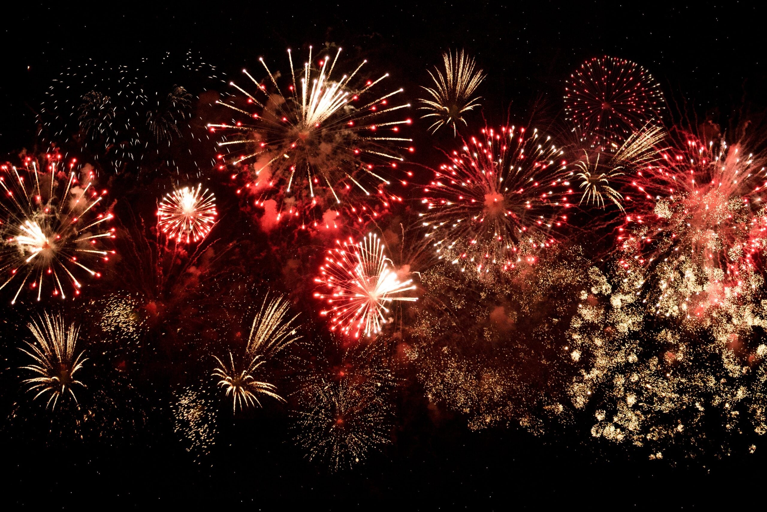 A vibrant display of fireworks lights up the night sky with bursts of red, gold, and white. Multiple patterns and shapes create a dazzling, festive scene against the dark background.