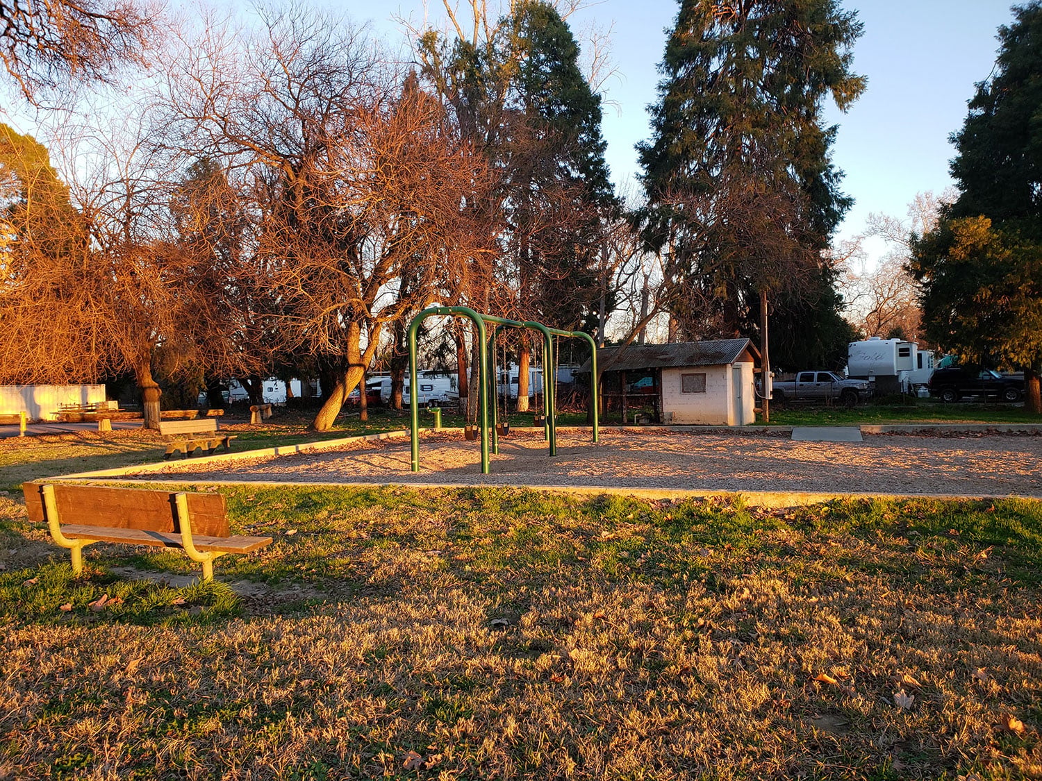 A small playground with swings surrounded by trees in a park. The ground is covered with sand, and a wooden bench is in the foreground. The sunlight casts long shadows, giving a warm tone to the scene.