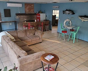 A cozy room with a brown sectional sofa on tiled floors, a small round table with green chairs, a kitchenette with red appliances in the background, and decorative fish mounted on blue-painted walls.