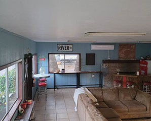 A cozy living room with blue walls, featuring a brown sectional sofa, bar stools by the window, a sign that reads "RIVER," and a small countertop with a vintage Coca-Cola dispenser in the corner.