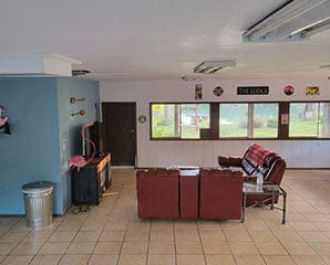 A spacious room with tiled floors features a wall-mounted TV, a red couch, and a small coffee table. Decorations include wall signs and musical instruments. Large windows provide a view of a garden outside. A pink flamingo statue stands by the TV.