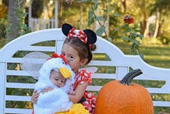 A young girl in a Minnie Mouse costume holds a baby dressed in a chicken outfit. They are sitting on a white bench in a garden, next to a large pumpkin.