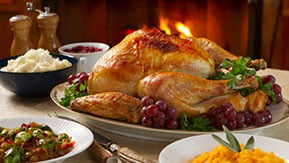A roasted turkey on a platter garnished with parsley and grapes, surrounded by side dishes including mashed potatoes, cranberry sauce, mixed vegetables, and a bowl of mashed sweet potatoes. A warm fire glows in the background.
