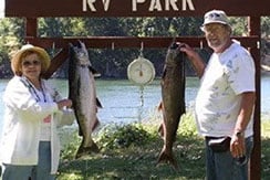 A woman and a man stand in front of a sign that says "RV Park," each holding a large fish hanging from a scale. They are outdoors near a body of water with trees in the background.