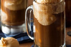 A frosty glass mug filled with root beer float, topped with scoops of vanilla ice cream. Another mug is blurred in the background, and an extra scoop of ice cream sits on the wooden surface nearby.