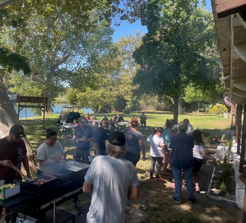 A group of people enjoying a barbecue in a sunny park. Several individuals are gathered around a grill, while others relax and socialize. Trees and a distant body of water are visible, creating a peaceful, outdoor atmosphere.