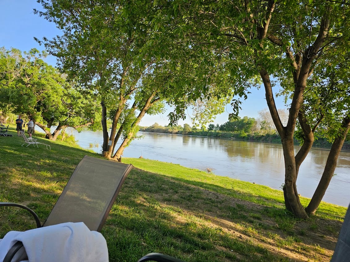 A serene riverside scene with lush green trees lining the bank. The river reflects the clear, blue sky. In the foreground, a lawn chair with a towel is visible, and a couple of people are enjoying the view on the grass.