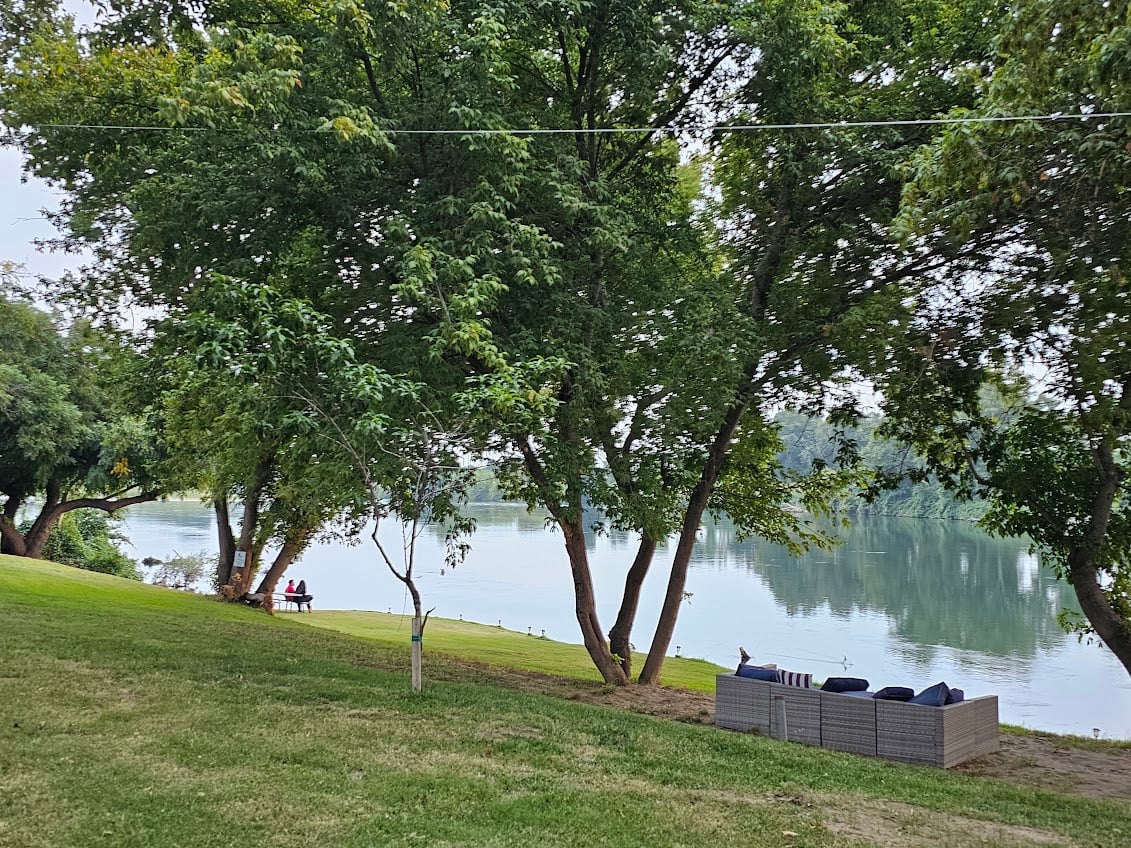 A peaceful lakeside scene with a couple sitting on a bench surrounded by lush green trees. A cozy outdoor couch is nearby, offering a relaxing spot to enjoy the tranquil view of the calm water under a cloudy sky.
