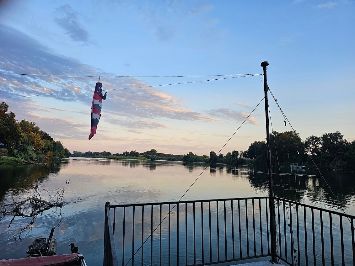 A serene riverside view with a docked boat in the foreground. A windsock flutters on a pole, and trees line both sides of the calm water under a partly cloudy sky at sunset.