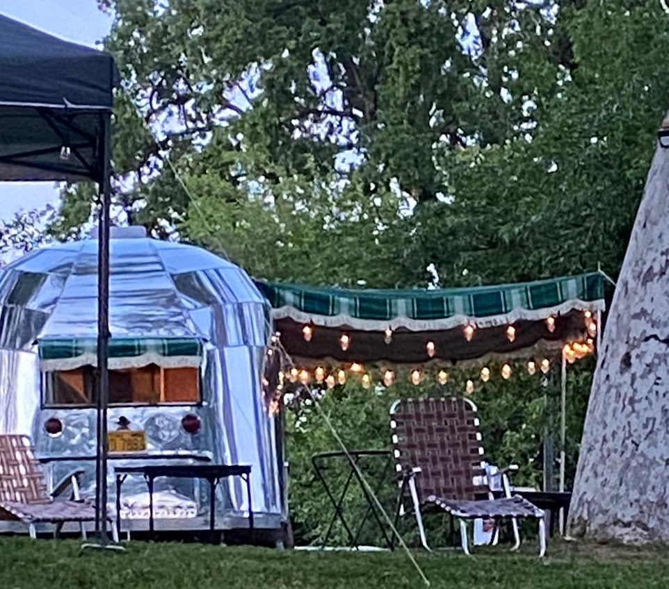 A silver camper trailer is parked near a canopy with string lights and two woven lounge chairs, set in a grassy area with trees in the background.