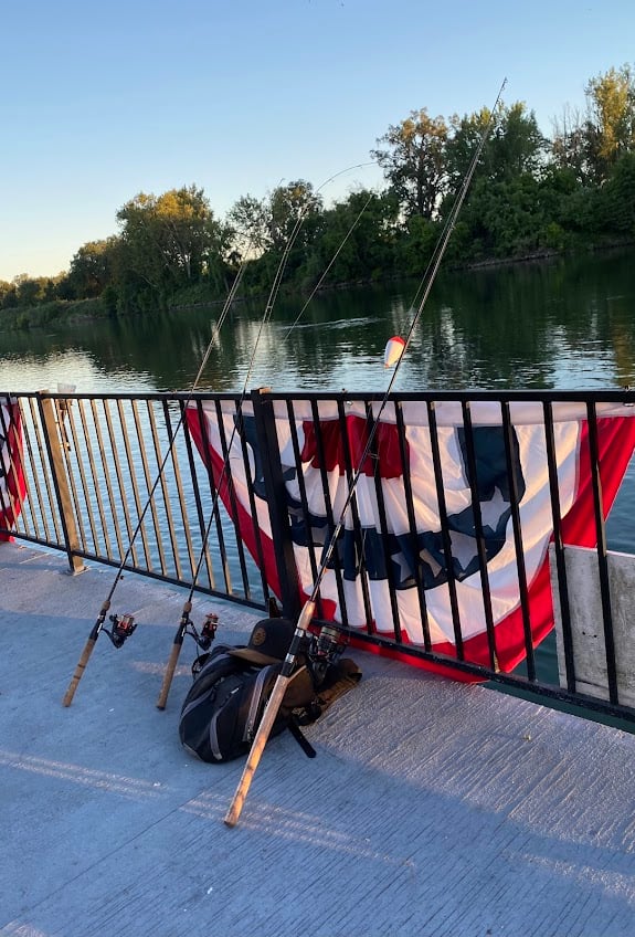 Three fishing rods are propped against a black railing overlooking a calm river. A large red, white, and blue decorative banner hangs on the railing. A black backpack sits next to the rods. Trees are visible in the background under a clear sky.