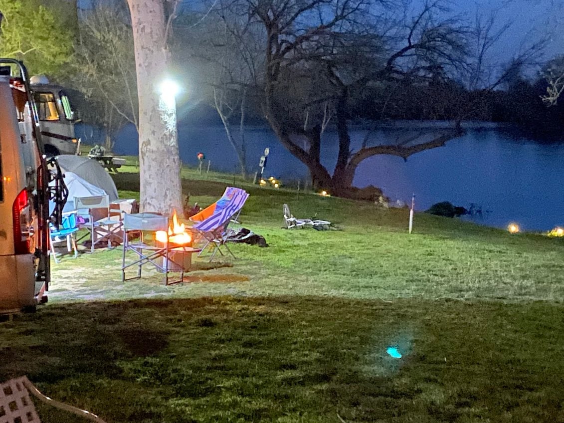 A nighttime camping scene by a lake, featuring a lit campfire surrounded by empty chairs. A tent is pitched nearby under a large tree, with an RV parked to the side. Soft outdoor lights illuminate the campsite.