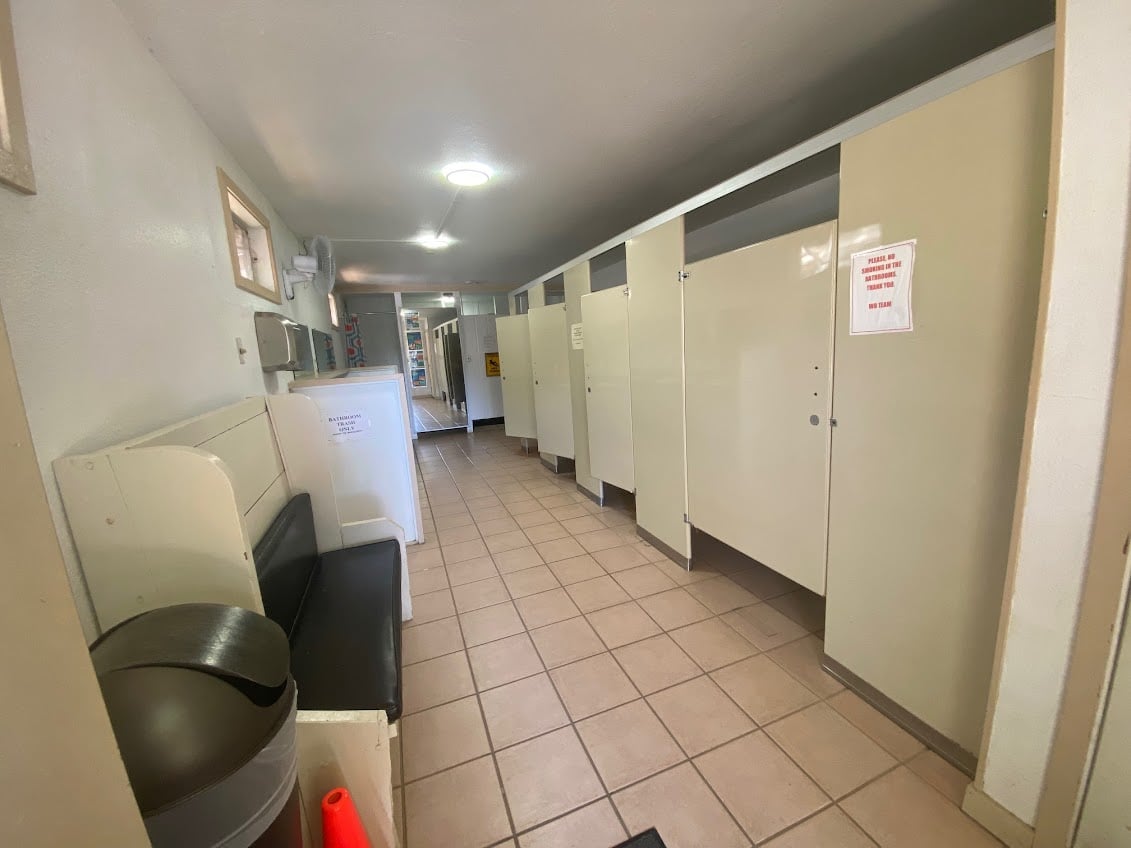 A hallway with tiled flooring and a row of beige restroom stalls on the right. A bench and a trash can are on the left side. The lighting is bright, and there are signs on some stall doors.