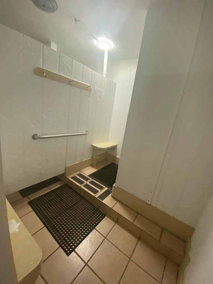 A narrow bathroom with a tiled floor and a shower area. The shower has wooden steps leading up, a grab bar on the side wall, and a small window near the ceiling. Black rubber mats are placed outside the shower area.