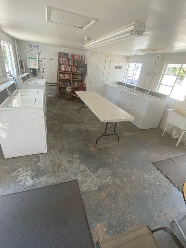 A laundry room with several washing machines and dryers lining the walls. A long folding table is in the center of the room. A bookshelf filled with books is against the far wall, and there are large windows bringing in natural light.