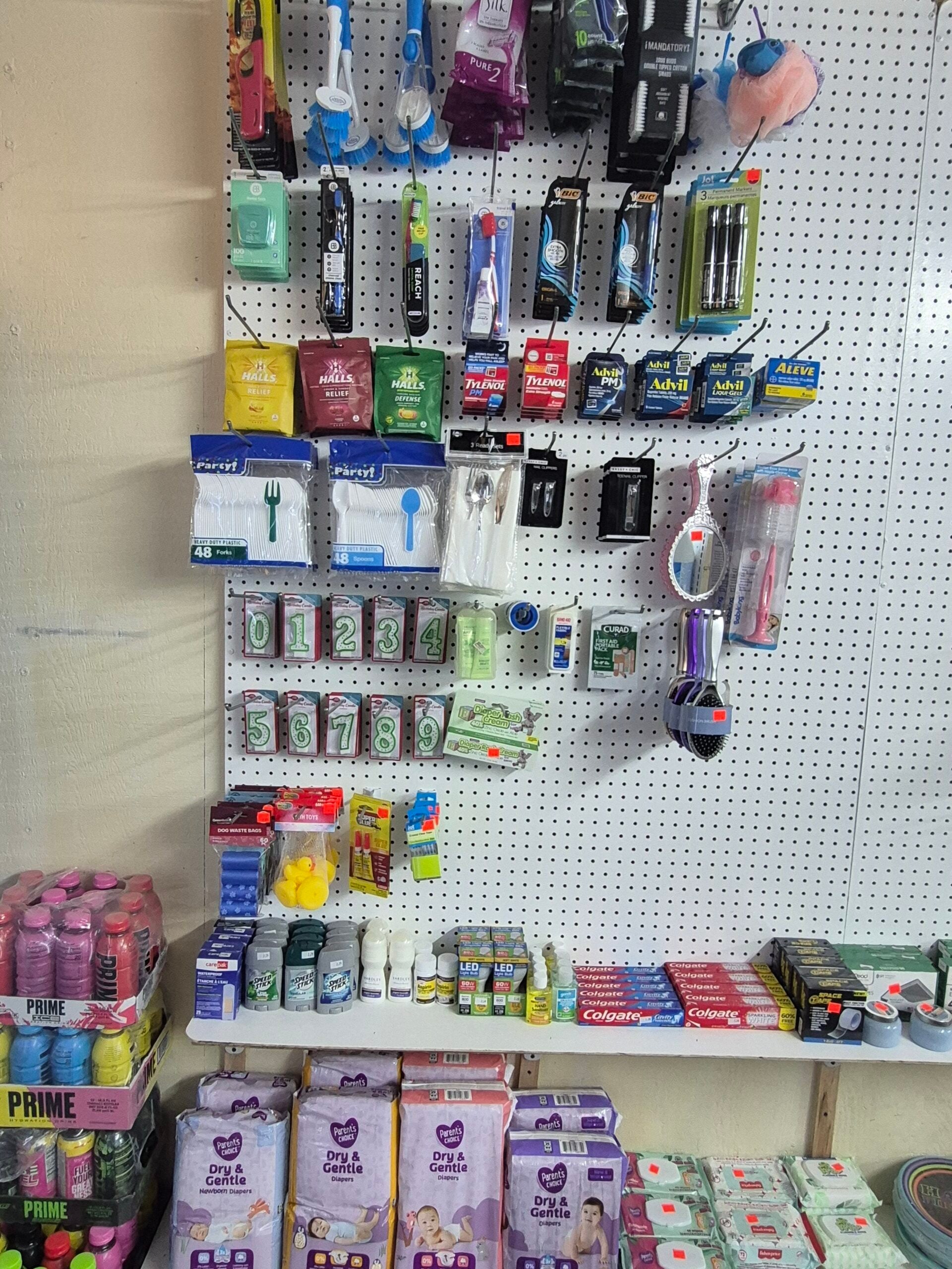 A store display with dental hygiene products, razors, bath sponges, and household items on a pegboard wall. Below, baby formula, diapers, canned drinks, and toothpaste are arranged on shelves and boxes.