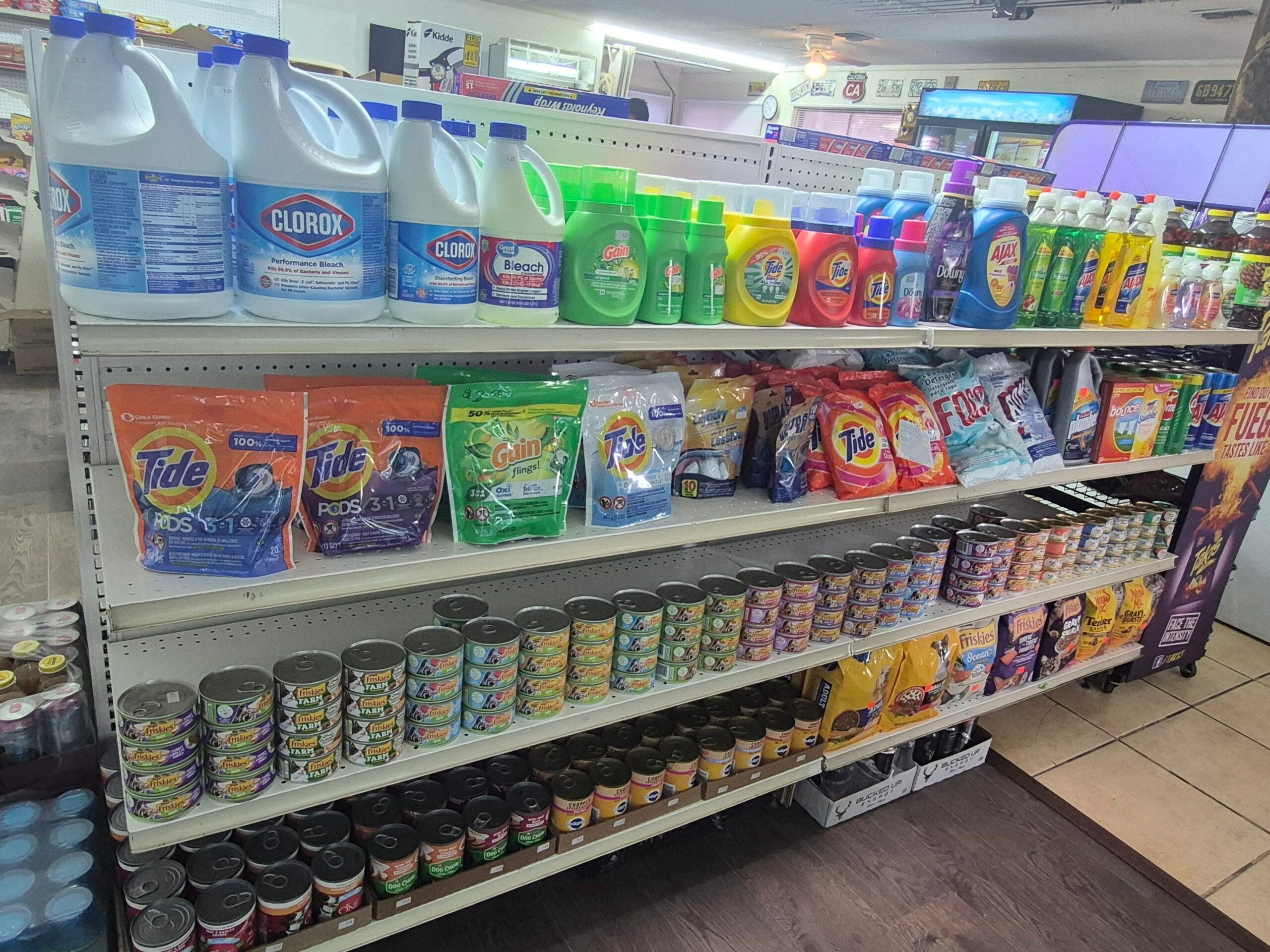 Shelves in a store stocked with laundry detergents, bleach bottles, dish soaps, canned cat food, and other cleaning and household products. The shelves are organized and fully packed.