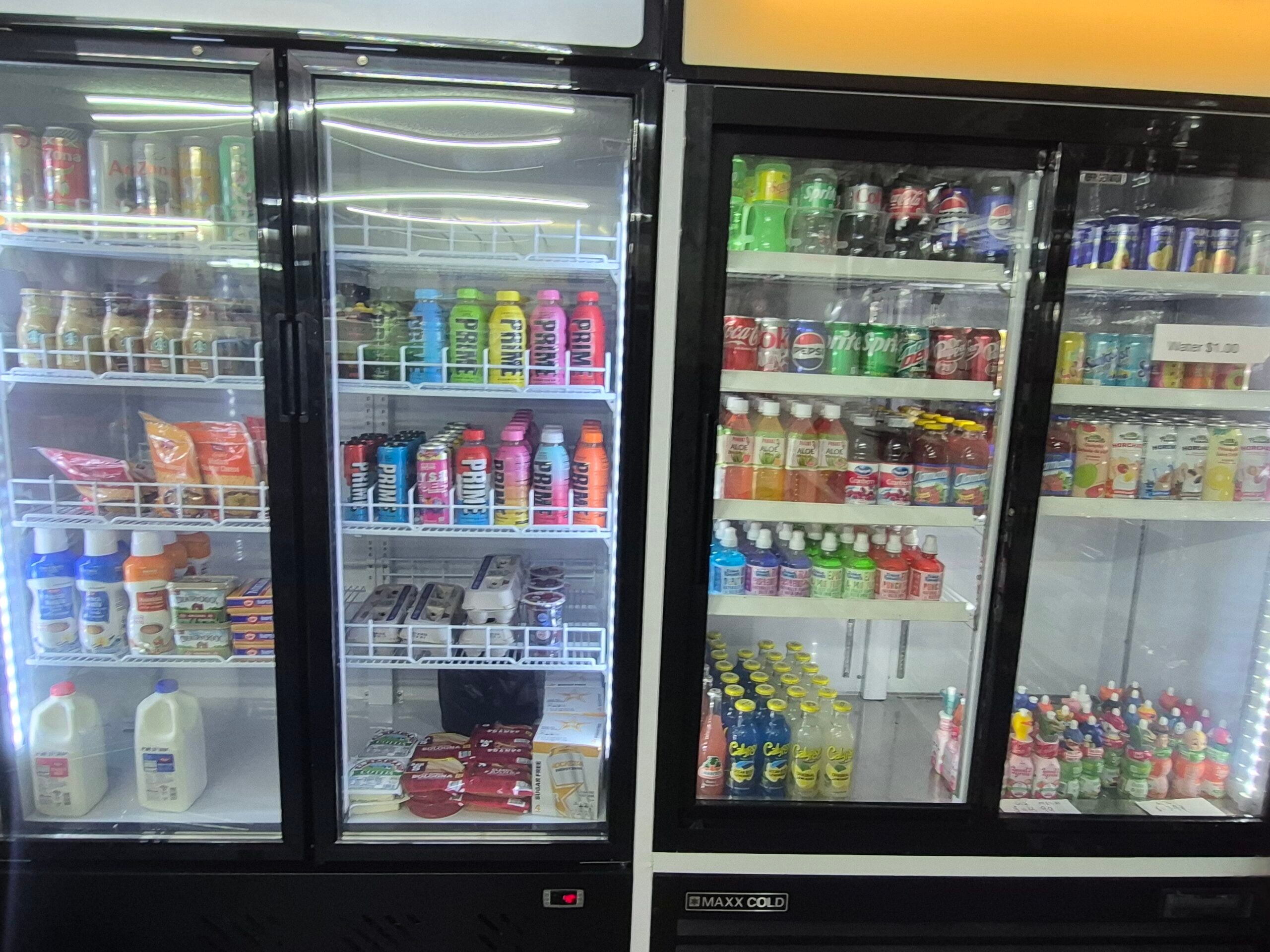 Refrigerator cases in a store stocked with various bottled drinks, sodas, energy drinks, dairy products like milk, and packaged snacks. Shelves are organized, with drinks and snacks visible behind glass doors.