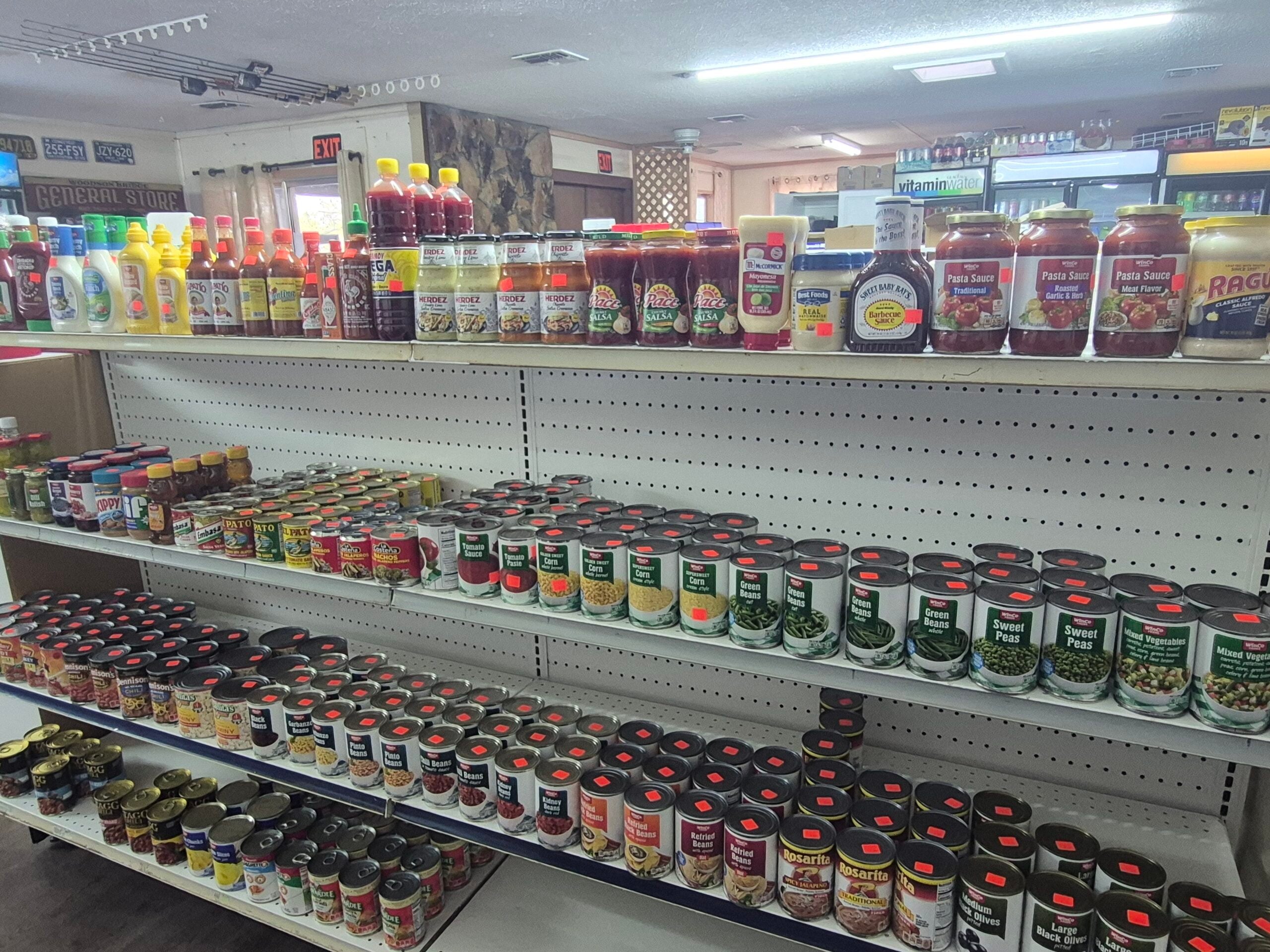 Shelves in a grocery store stocked with various canned vegetables, sauces, and condiments. Bright lights illuminate the products, which are organized in neat rows with red price stickers visible on many items.