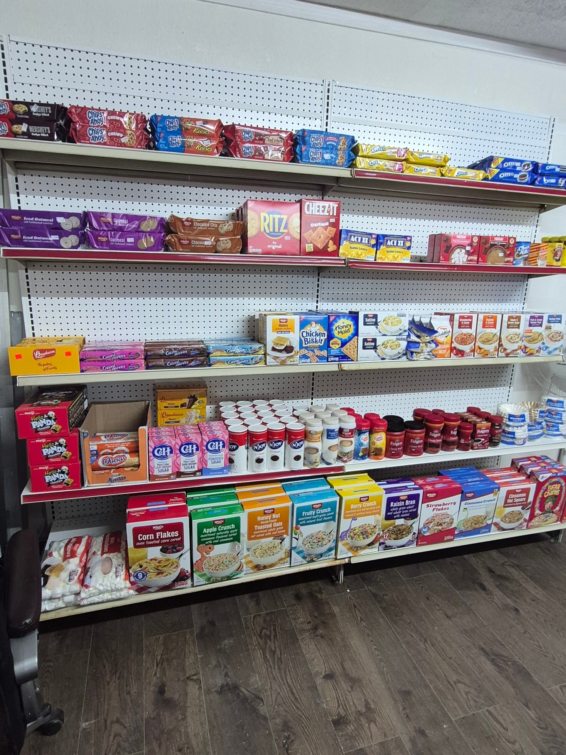 Shelves stocked with various packaged food items, including cereals, crackers, cookies, peanut butter, jelly, sugar, salt, and baking mixes. The floor is wooden, and the products are neatly arranged.